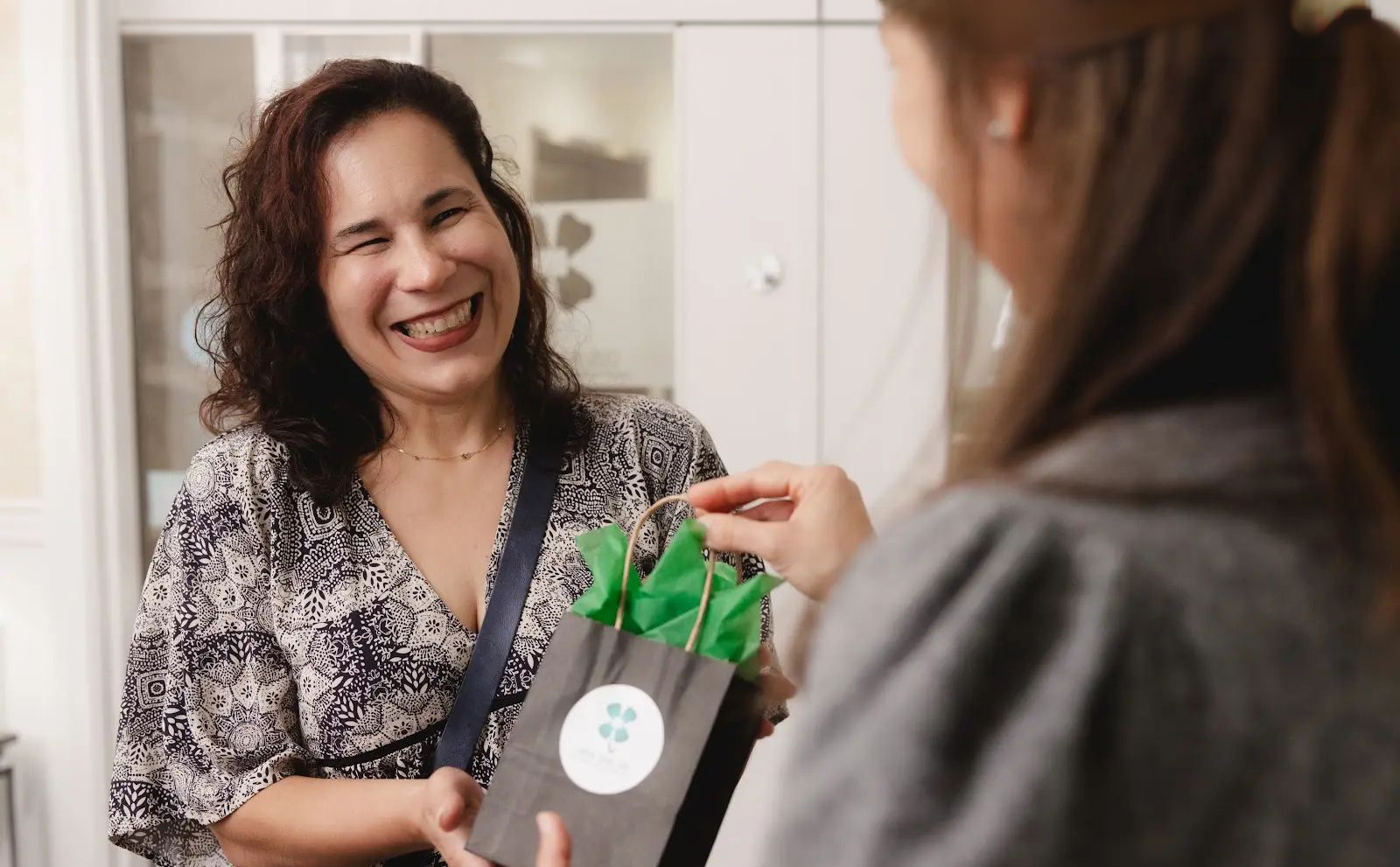 A smiling woman with curly dark hair, wearing a patterned dress, receives a small gray gift bag with green tissue paper from another person.