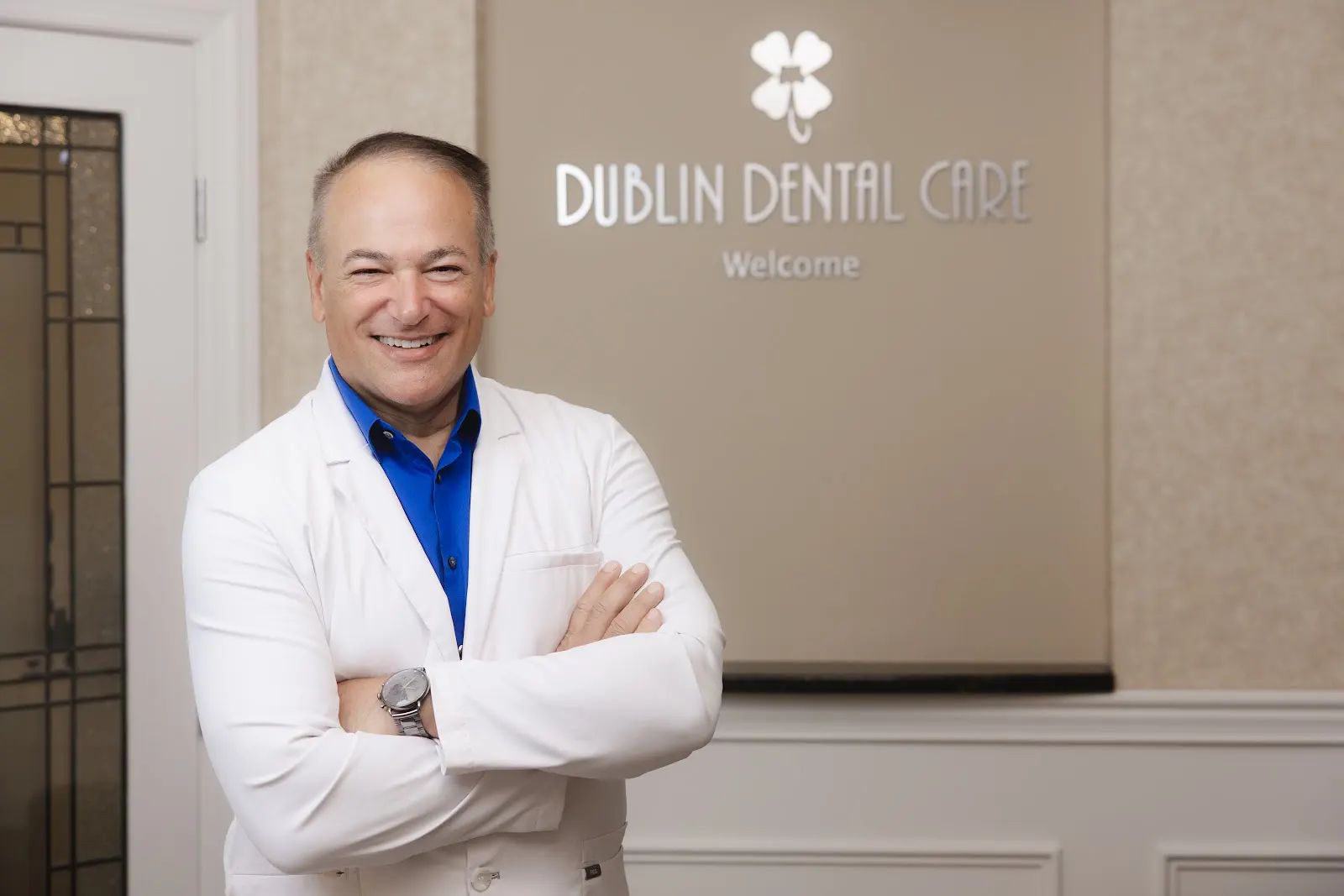 Smiling male dentist in a white coat and blue shirt standing with arms crossed in front of a wall sign that reads 'Dublin Dental Care Welcome'.