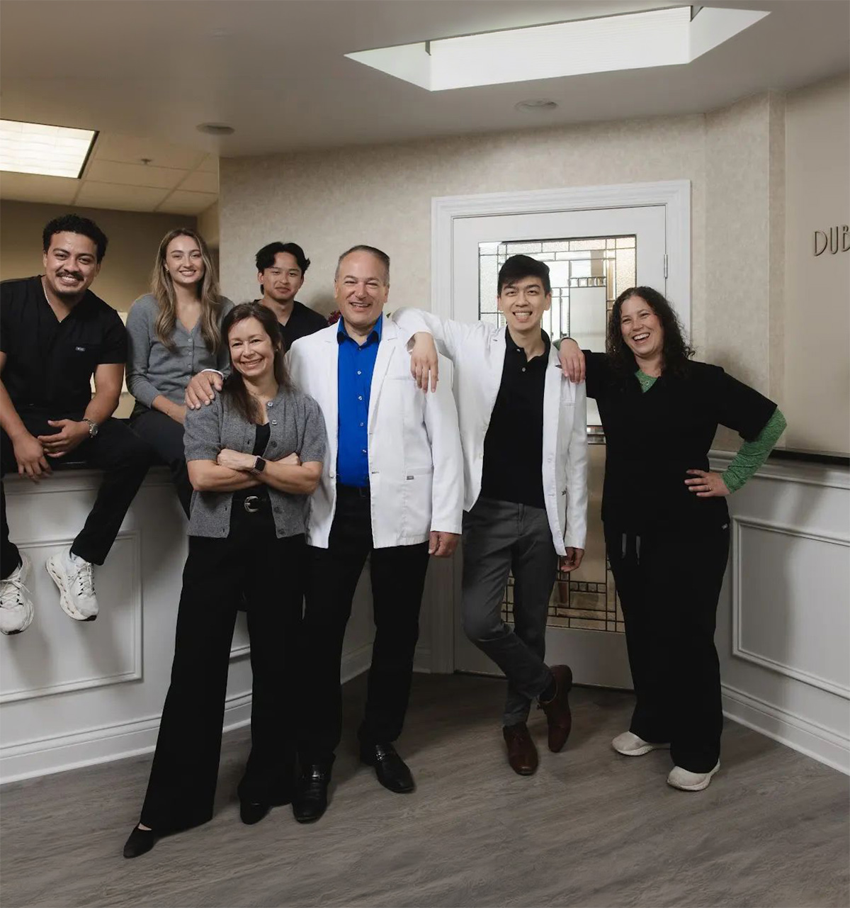 Group of seven smiling medical professionals in a clinic reception area, including two men in white coats standing center, others in black scrubs or casual wear, posing warmly together.