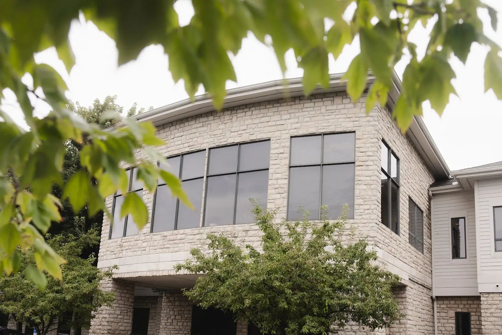 Stone building with large dark windows on the upper floor and green tree branches in the foreground.