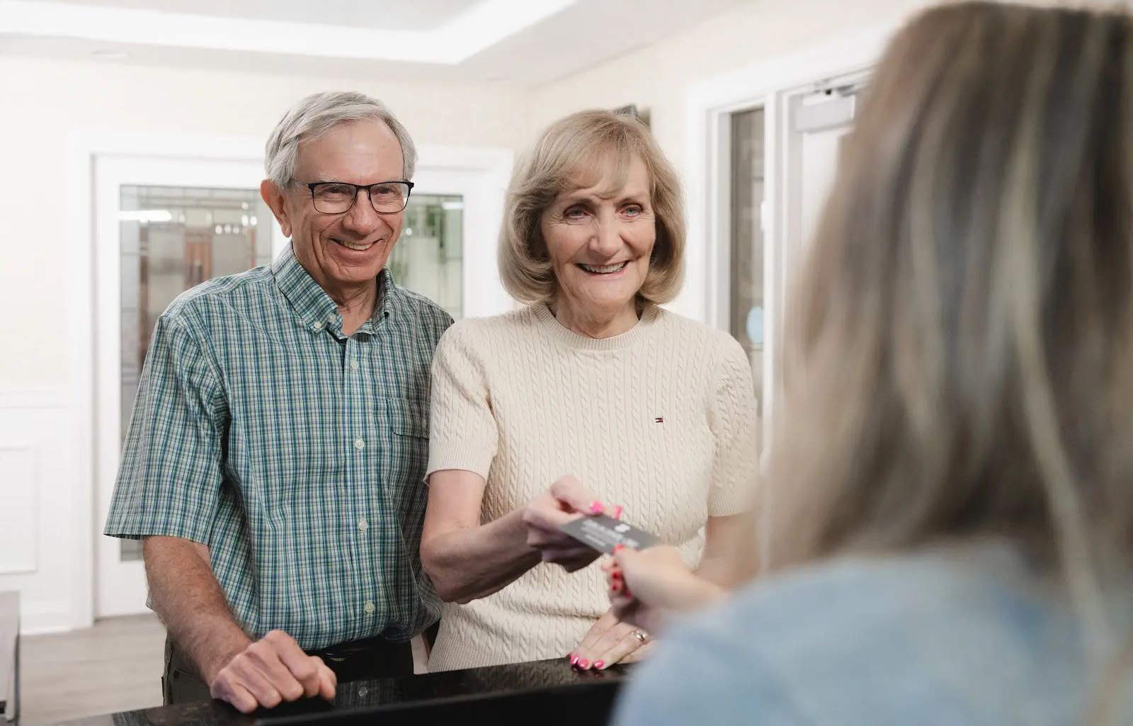 Smiling elderly couple standing at a reception desk, with the woman handing a card to a receptionist whose back is to the camera.