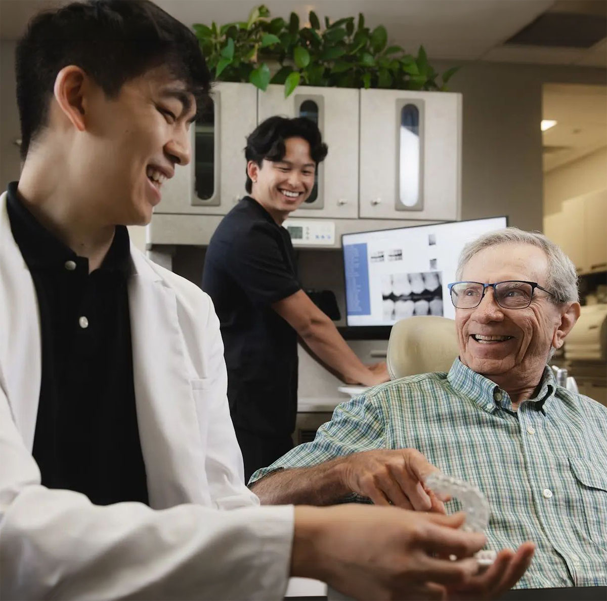 An older man sitting in a dental chair smiling and holding a clear dental aligner while a young male dentist in a white coat smiles back at him and a second young male staff member stands in the background near a computer showing dental X-rays.