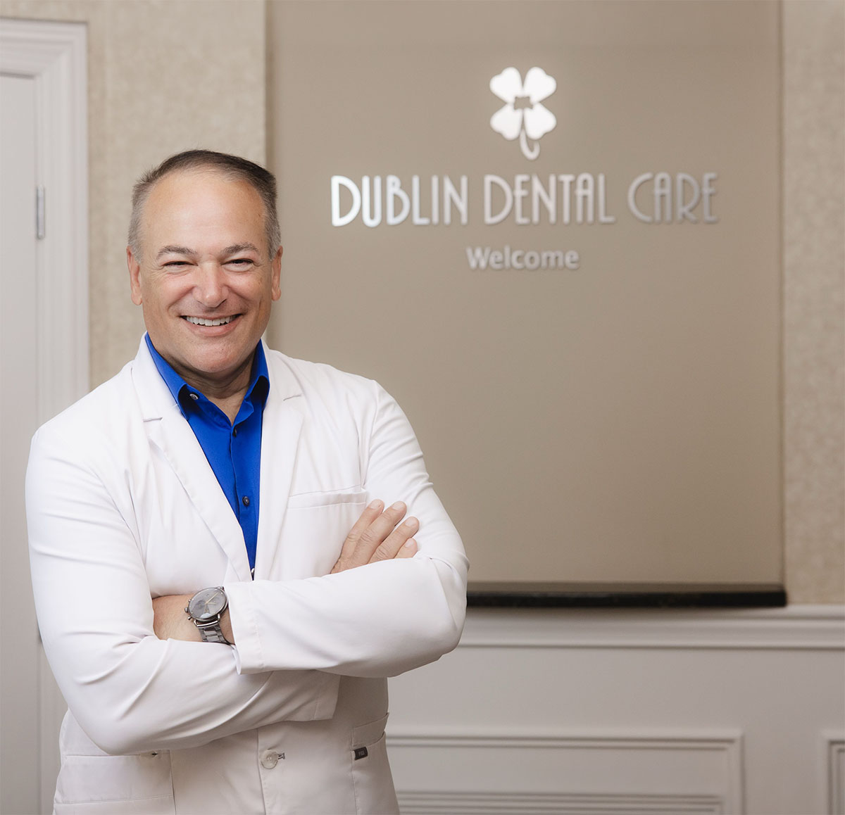 Smiling male dentist in a white coat and blue shirt standing with arms crossed in front of a wall sign that reads 'Dublin Dental Care Welcome' with a four-leaf clover logo above it.