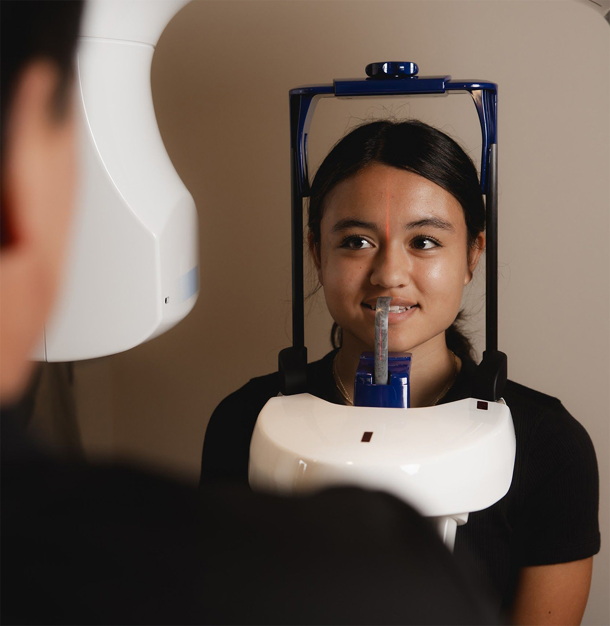 Young woman sitting in a dental imaging machine with a bite piece in her mouth and a red alignment laser on her forehead, preparing for a dental X-ray.