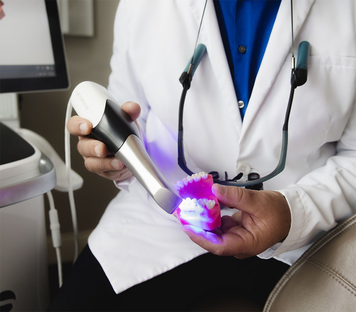 Dentist wearing a white coat holds a dental scanner emitting blue light over a pink dental model showing upper and lower teeth.