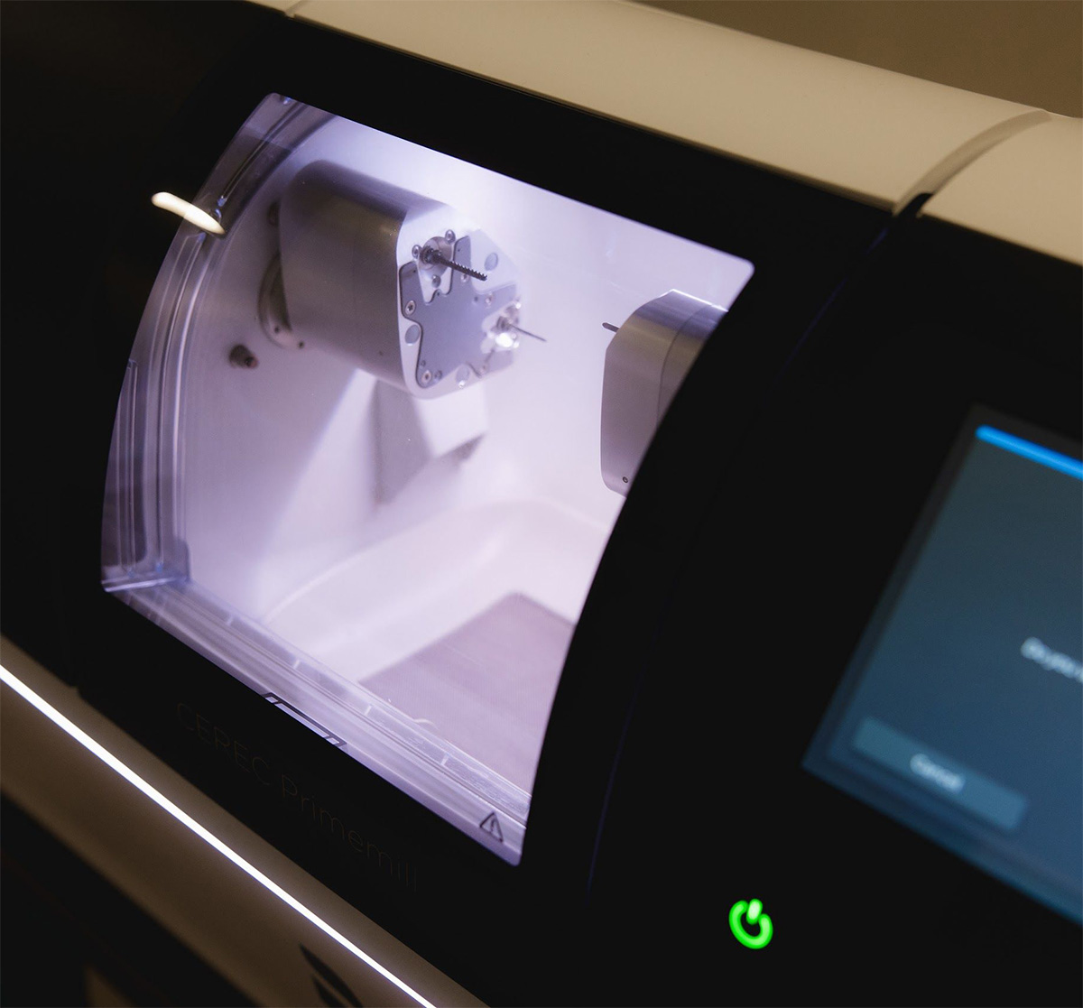 Close-up of a CEREC Primemill dental milling machine with a lit interior showing milling components and a touchscreen interface with a green power button.