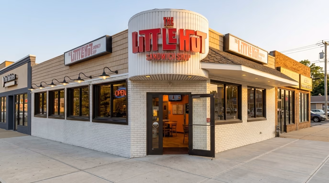 Corner view of 'The Little Hot Sandwich Shop' with white brick walls, large windows, and an open entrance door showing interior seating.