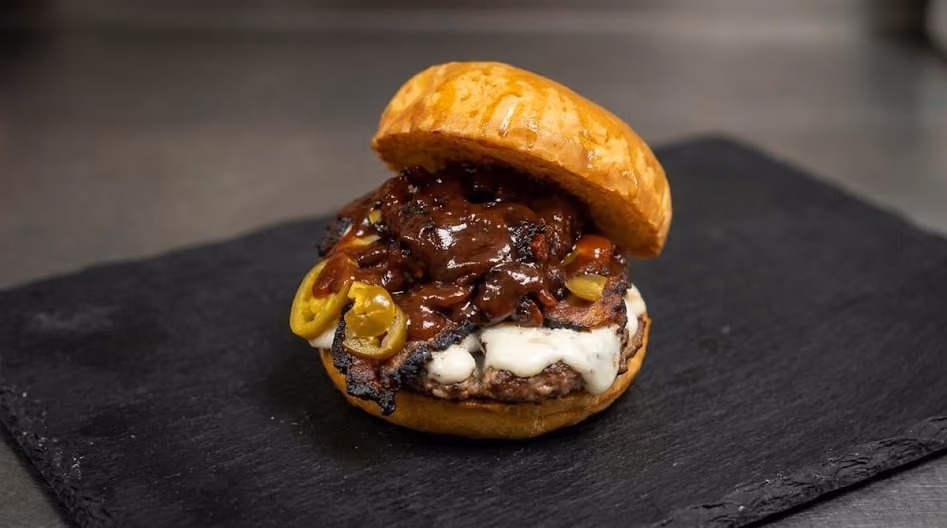 Burger with a toasted bun, melted white cheese, sliced jalapeños, thick slices of grilled meat, and a rich dark barbecue sauce, placed on a black slate surface.