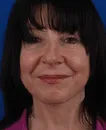 Close-up of a middle-aged woman with straight black hair and bangs smiling gently against a blue background.