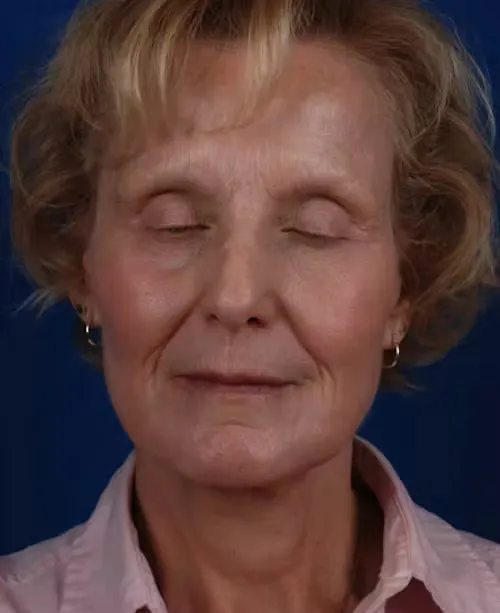 Close-up of an elderly woman with short curly hair, wearing small hoop earrings and a pink collared shirt, eyes closed against a dark blue background.