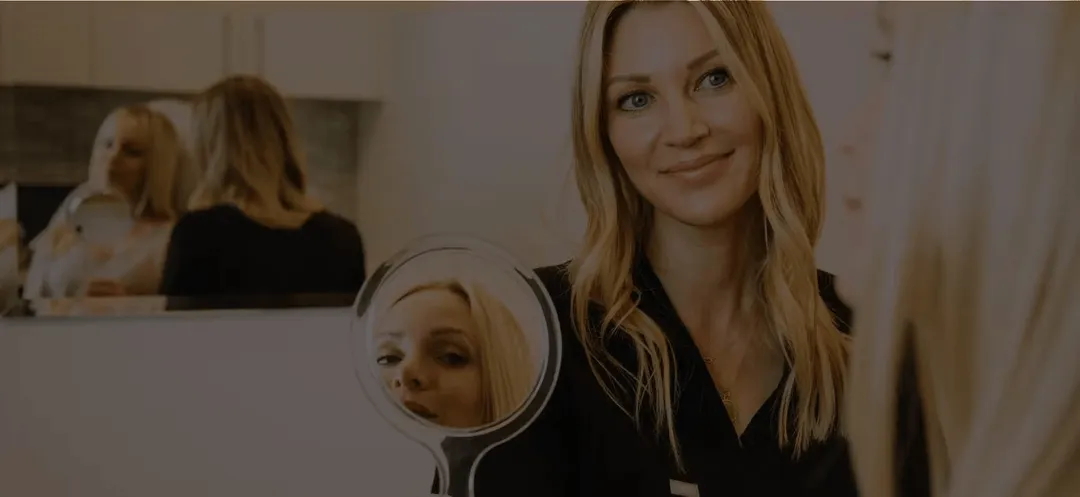 Smiling blonde woman with loose waves looking into a handheld mirror showing her reflection.