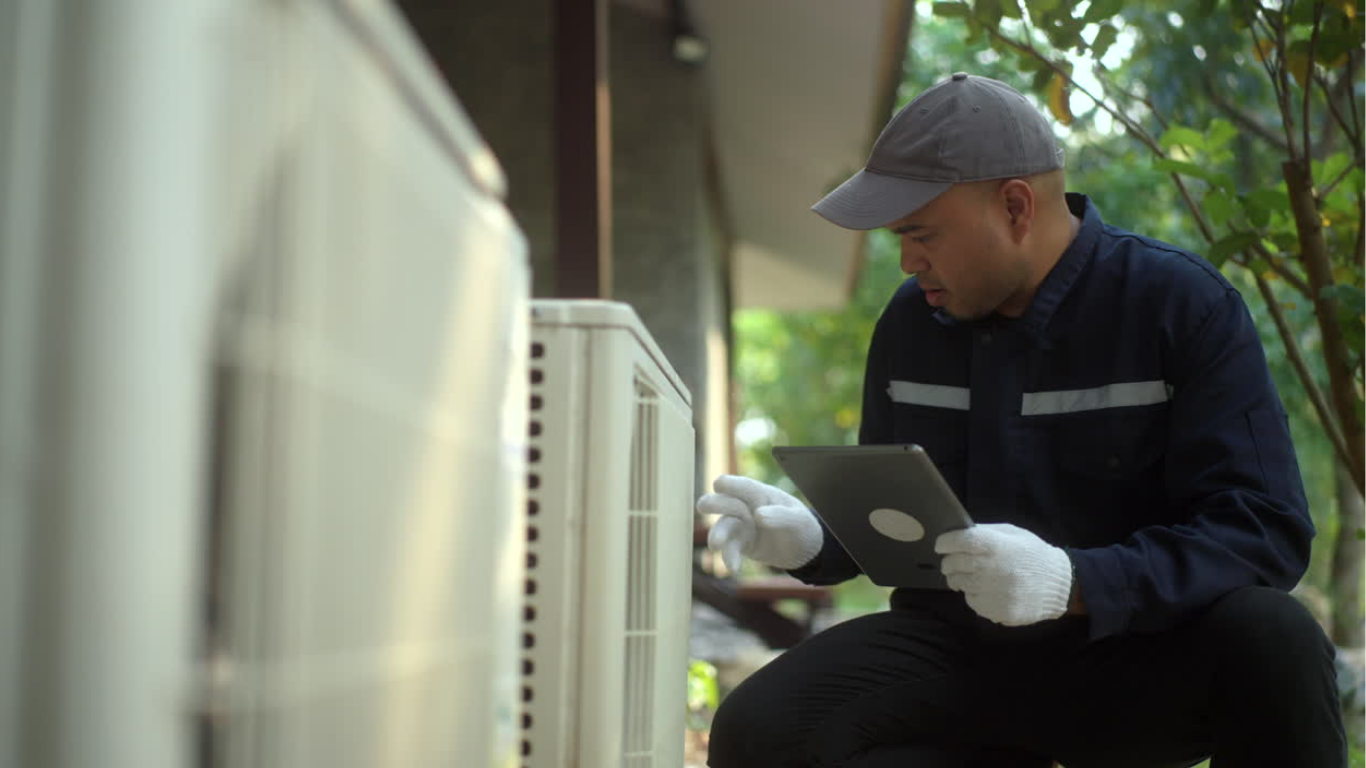 A technician wearing a gray cap, white gloves, and dark uniform inspects an outdoor air conditioning unit while holding a tablet.