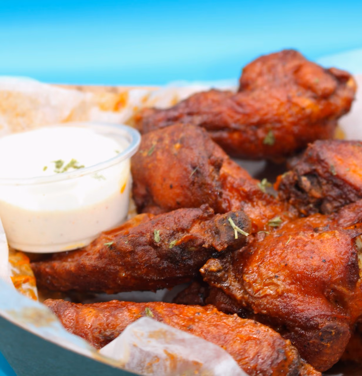 Close-up of crispy, seasoned chicken wings garnished with herbs in a tray with a cup of creamy dipping sauce.