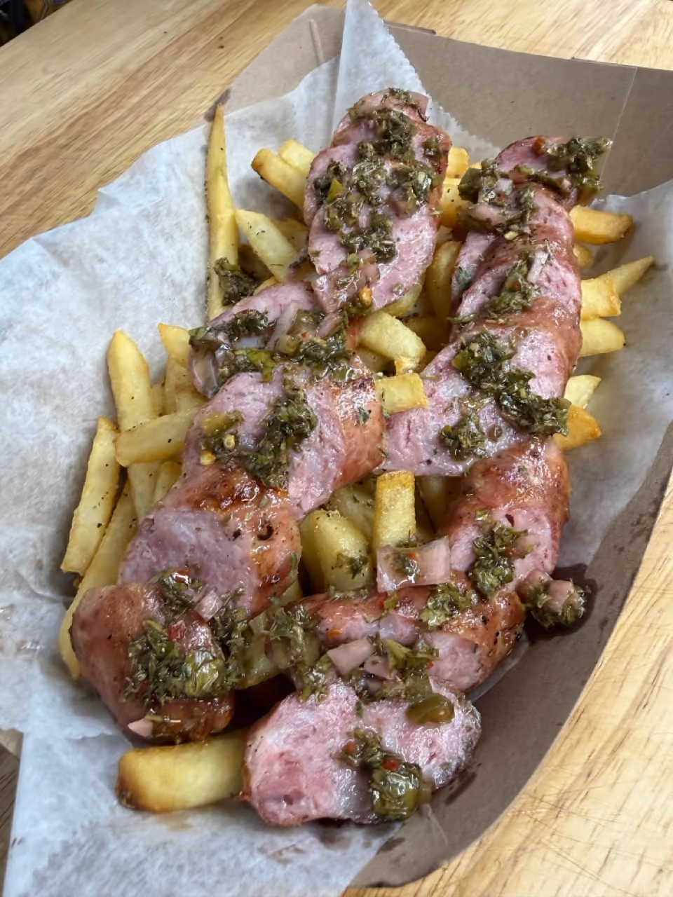 Sliced grilled sausage topped with chopped green herbs and spices served on a bed of golden French fries in a paper-lined cardboard tray.