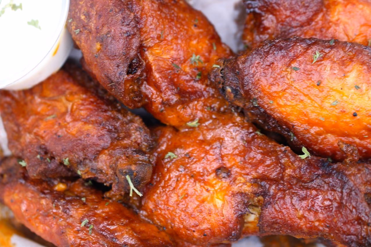 Close-up of several crispy, reddish-brown chicken wings garnished with small green herbs, with a portion of white dipping sauce visible on the side.