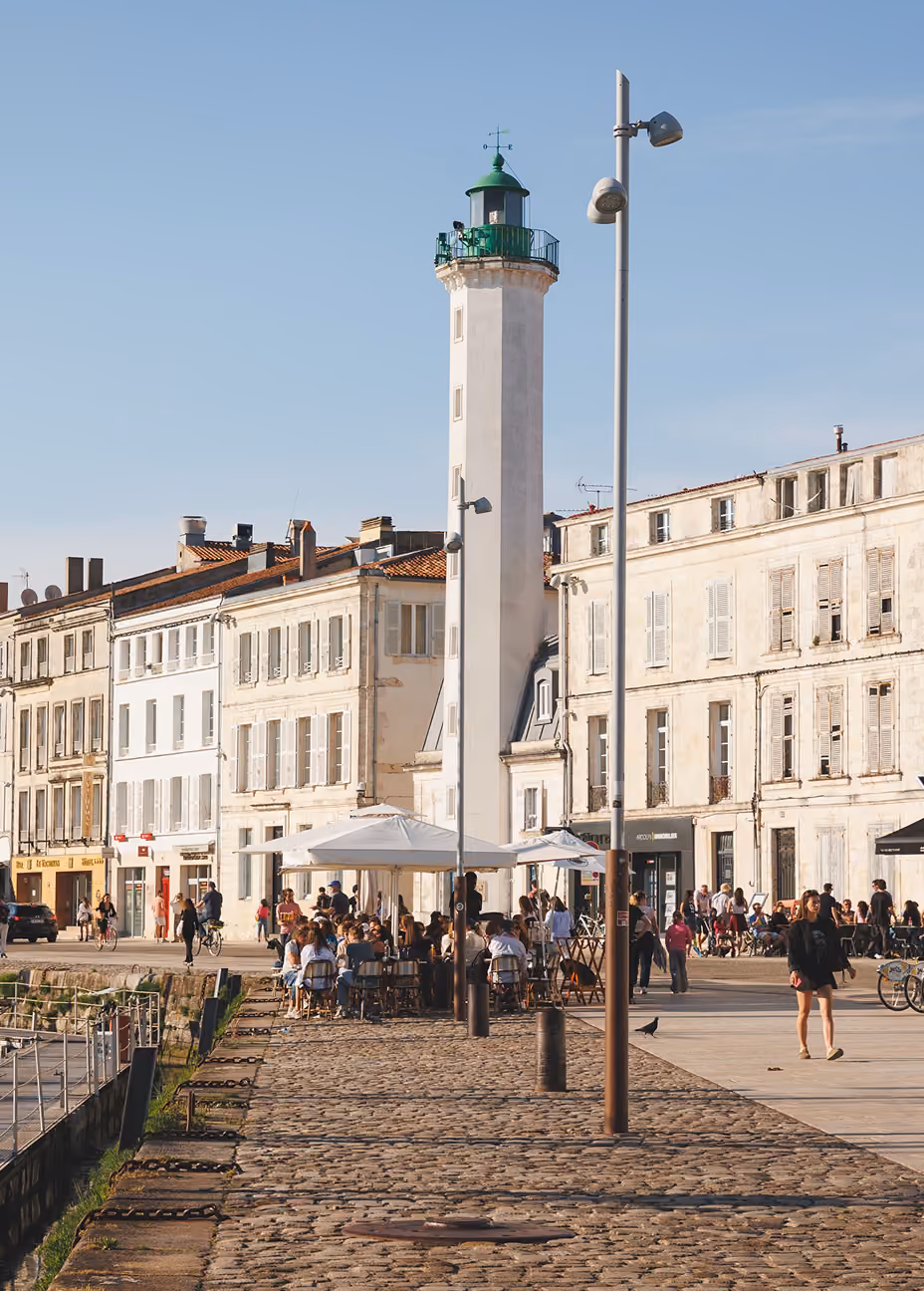 White lighthouse with green top by waterfront with people dining and walking on cobblestone path.