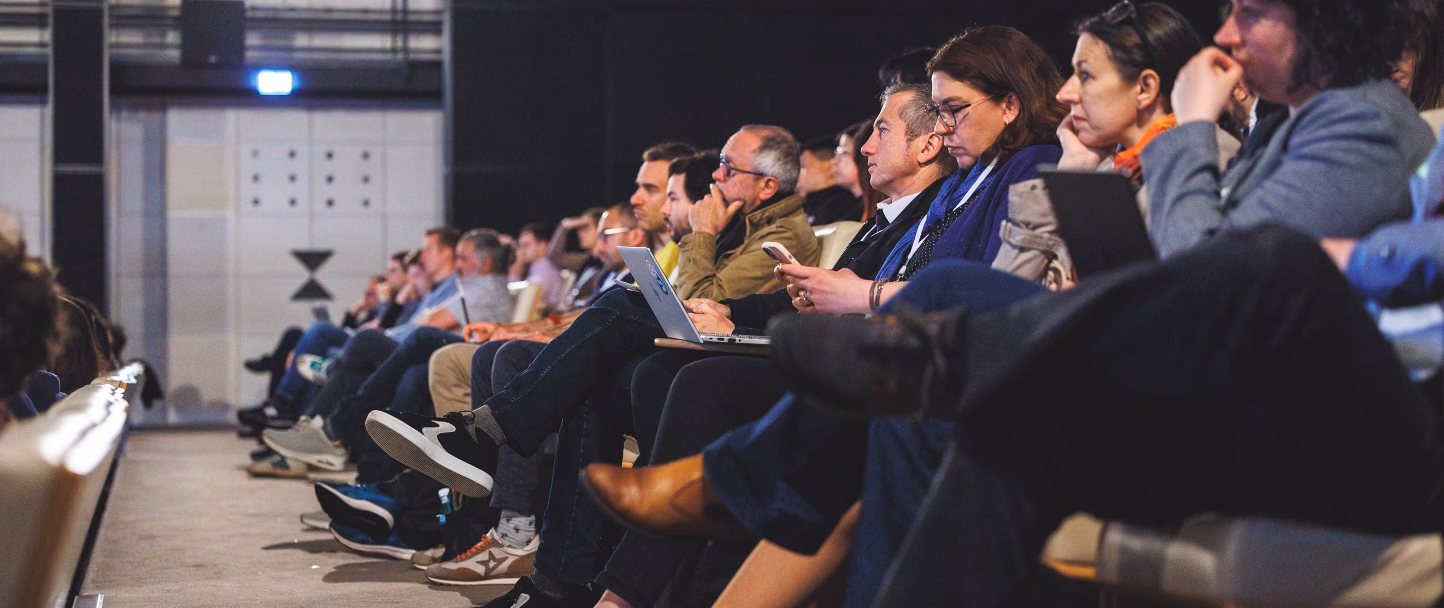 Audience sitting in a conference hall, some using laptops and phones, attentively engaged.