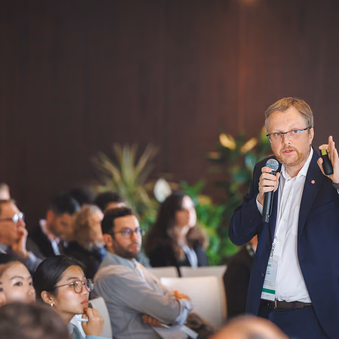 Man in glasses and suit speaking into a microphone at a conference with seated audience listening.