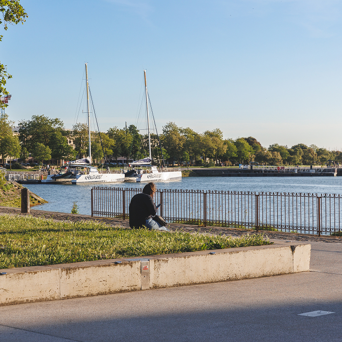 Person sitting on grass by waterfront with two sailboats docked nearby on a sunny day.