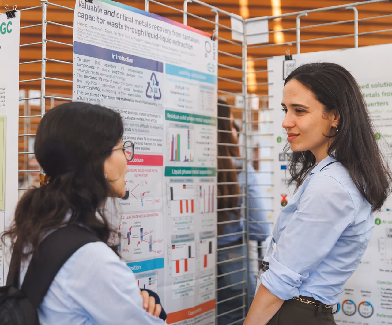 Two women discussing scientific posters displayed on a metal grid at a conference or exhibition.
