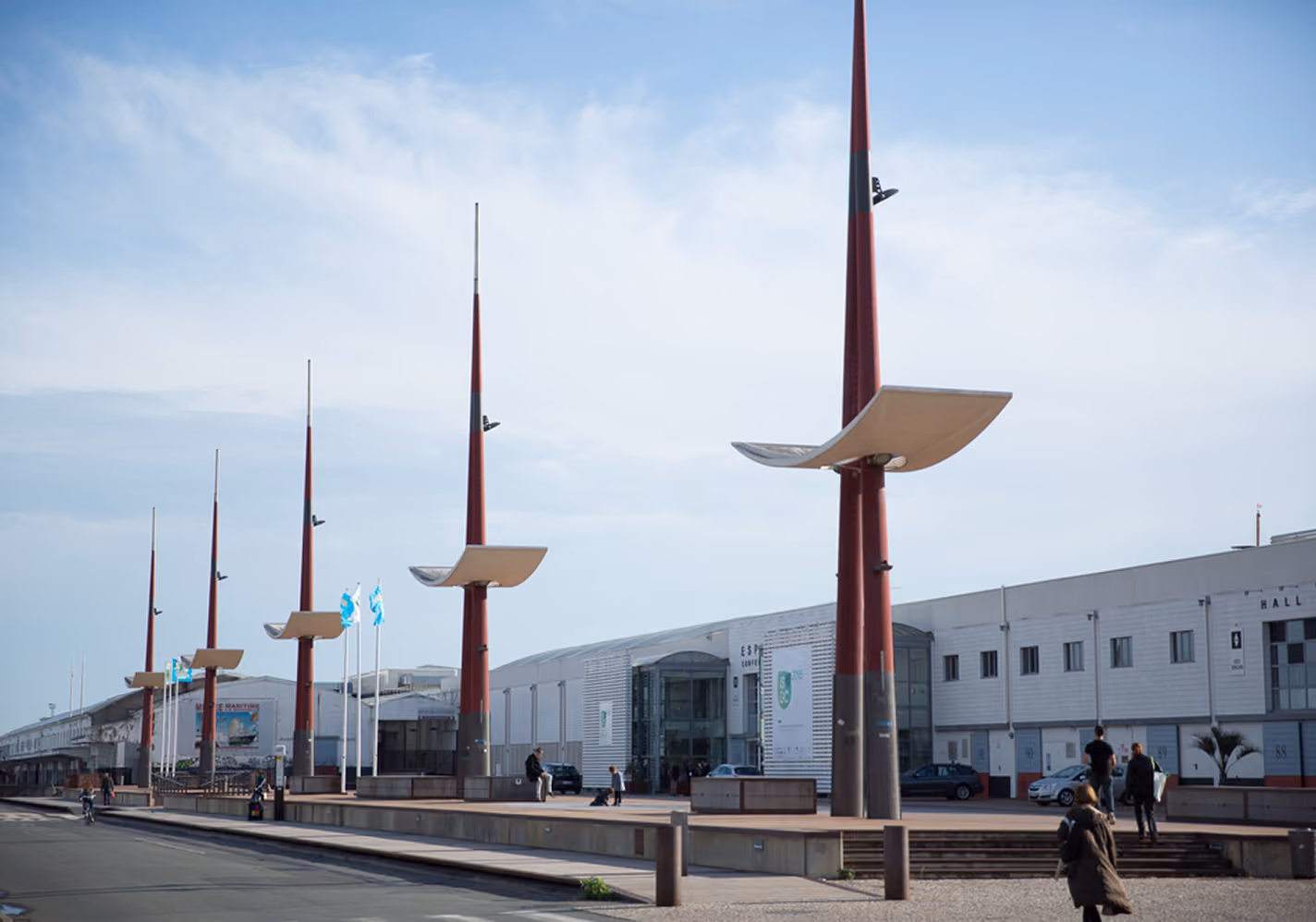 Row of tall red light poles with curved white fixtures along a wide walkway near industrial buildings.