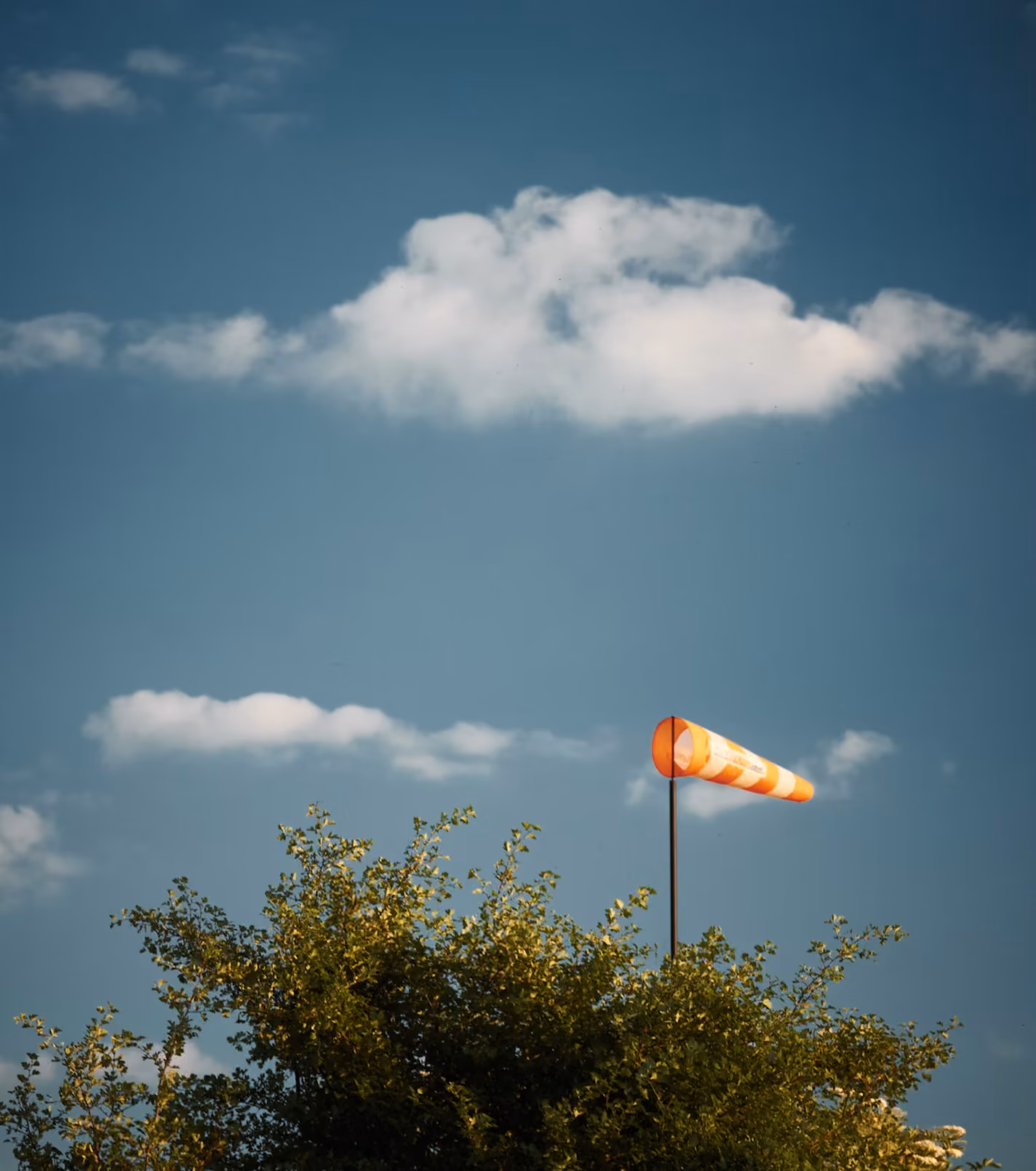Orange and white windsock above green bushes under a blue sky with scattered clouds.
