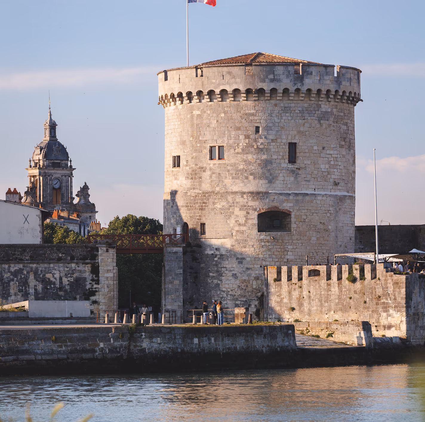 Stone tower with small windows and French flag near water with historic buildings in background.