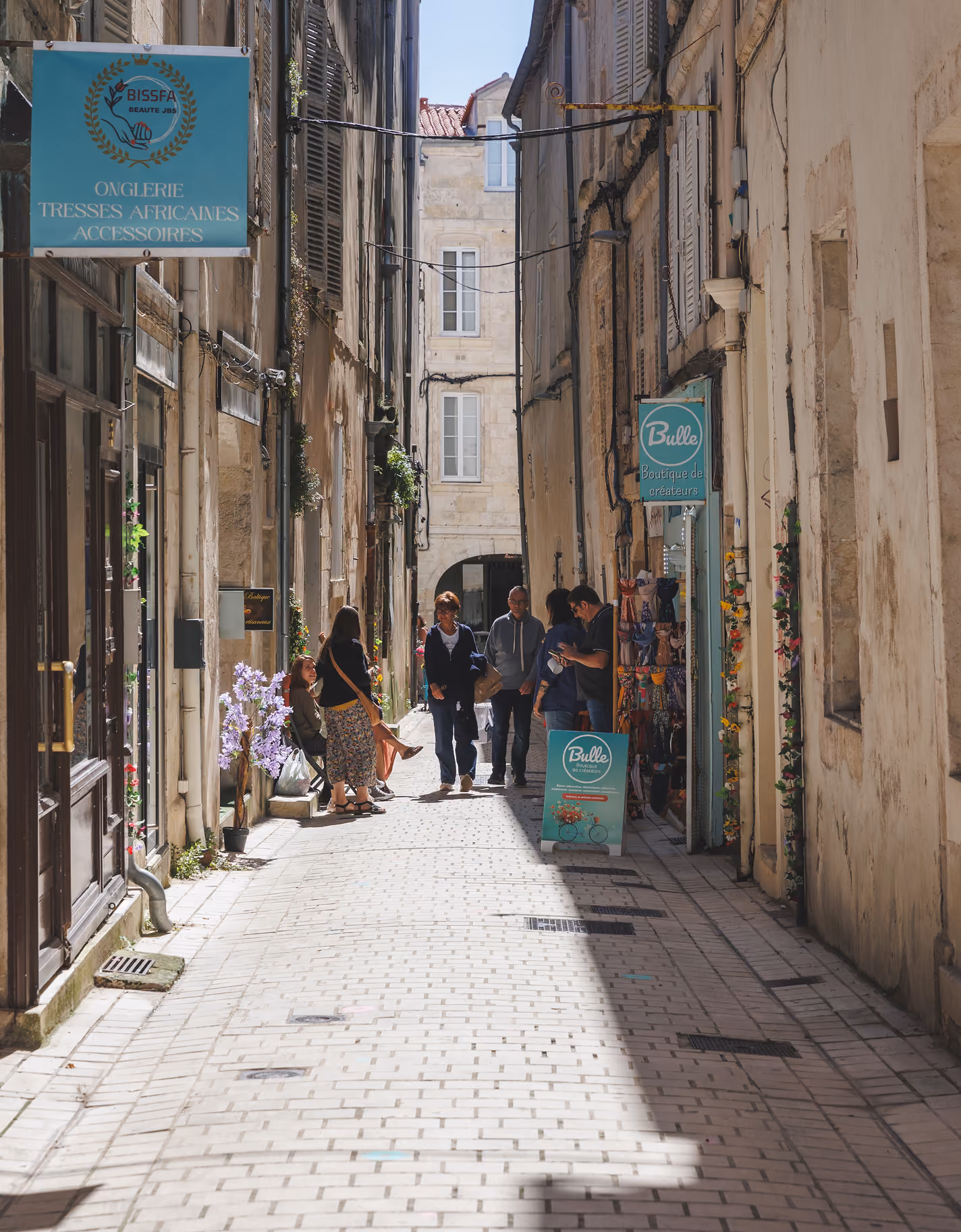 Narrow cobblestone alley flanked by tall, aged buildings with shuttered windows; pedestrians walk and stand near storefronts with signs, plants, and hanging items in soft sunlight.