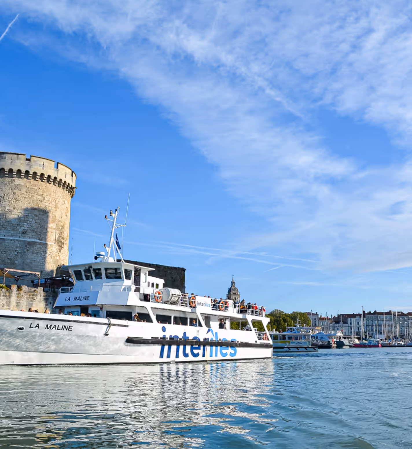 White passenger ferry named La Maline with interlines branding sailing on calm water near a historic stone tower under a blue sky with wispy clouds.