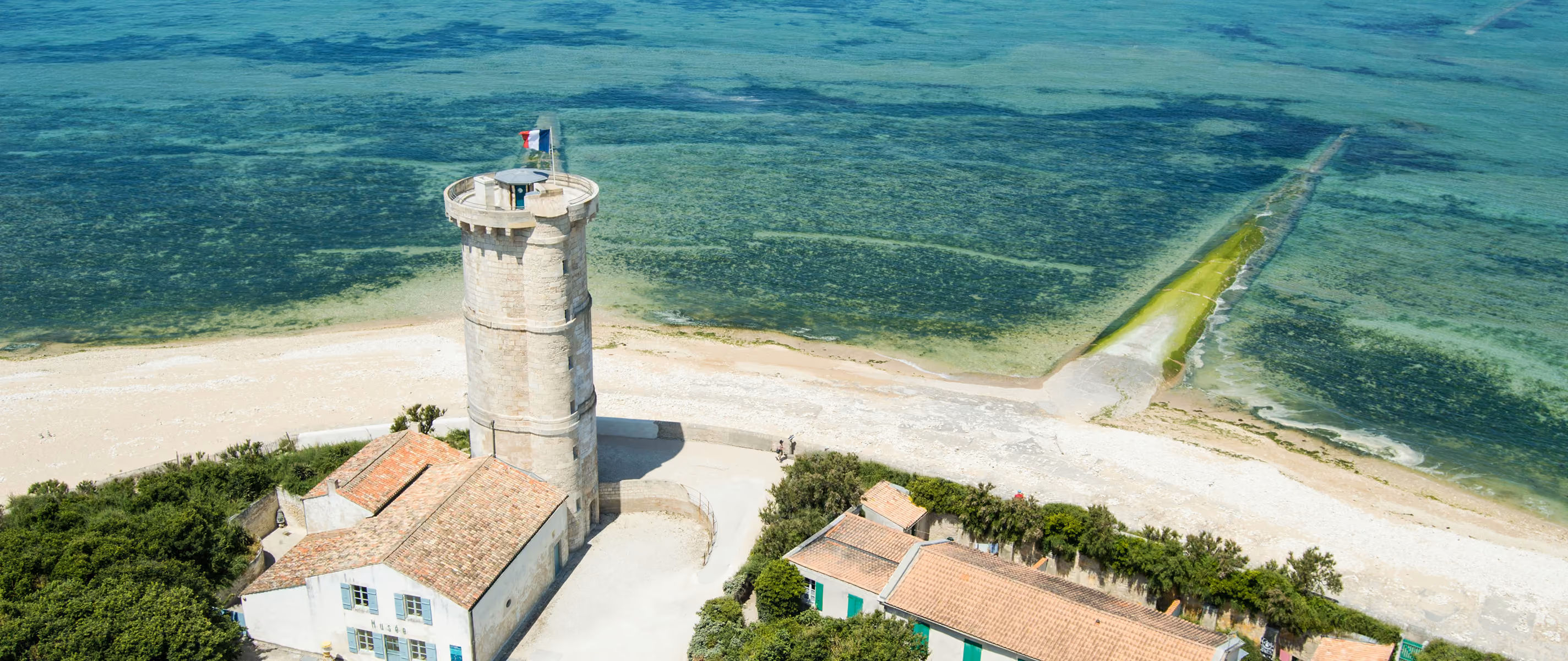 Aerial view of a round stone tower with a French flag on top, adjacent to buildings with tiled roofs, near a sandy shore and clear blue water.