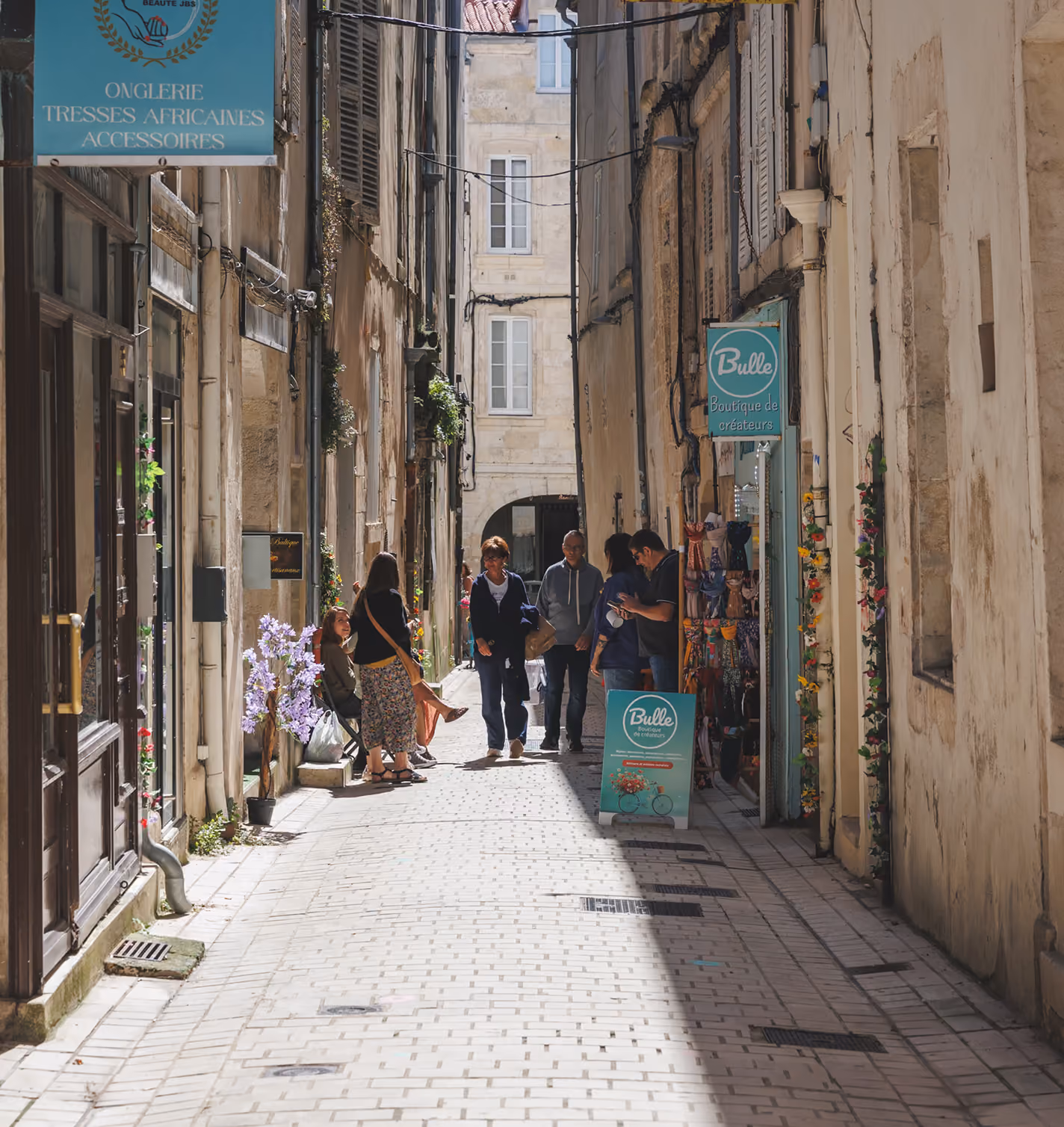 Sunlit narrow cobblestone street between tall old buildings with signs for African braids salon and 'Bulle' creator boutique, with groups of people walking and chatting.