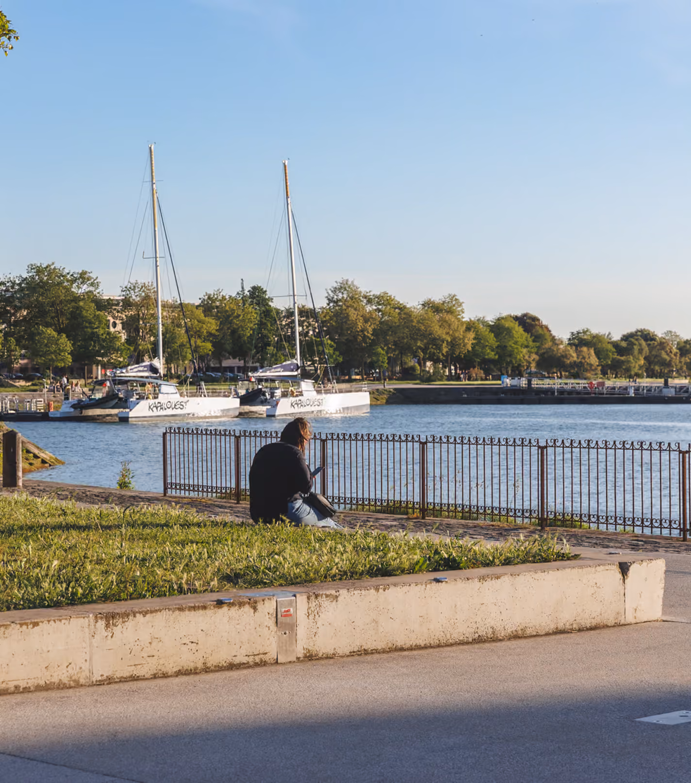 Person sitting on a ledge near a waterfront with a railing, facing a body of water where two sailboats are docked, with trees and clear blue sky in the background.