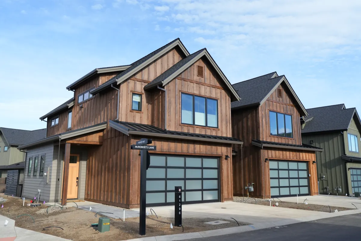 Two modern two-story houses with dark wood siding and large frosted glass garage doors on a residential street under a partly cloudy sky.