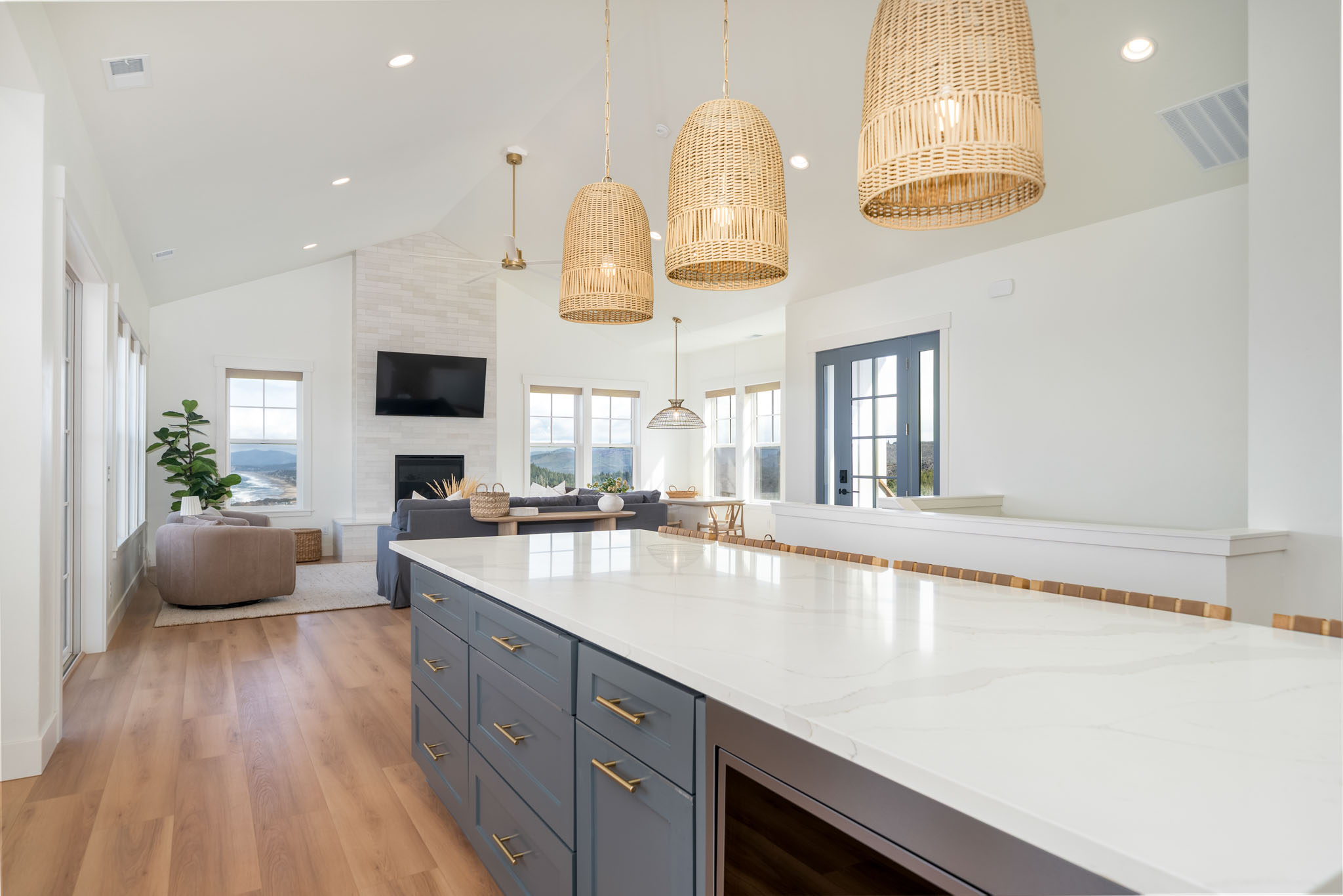 Bright, modern open-plan living space with a large white kitchen island with gray cabinets and brass handles, three woven pendant lights above it, a cozy seating area with a beige armchair and gray sofa, a fireplace with a mounted TV, and large windows showing a scenic landscape outside.