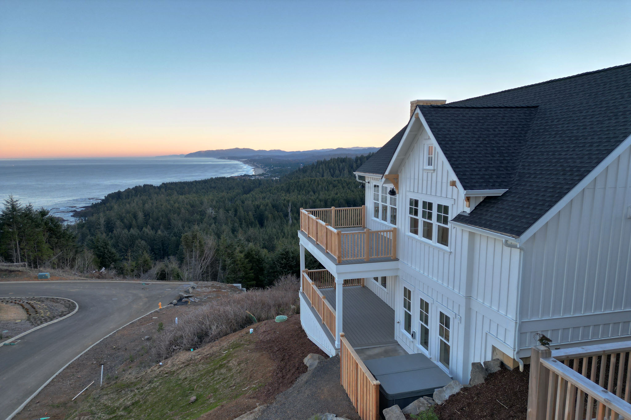 White house with black roof and wooden rail balconies overlooking a dense pine forest and a coastline at sunset.