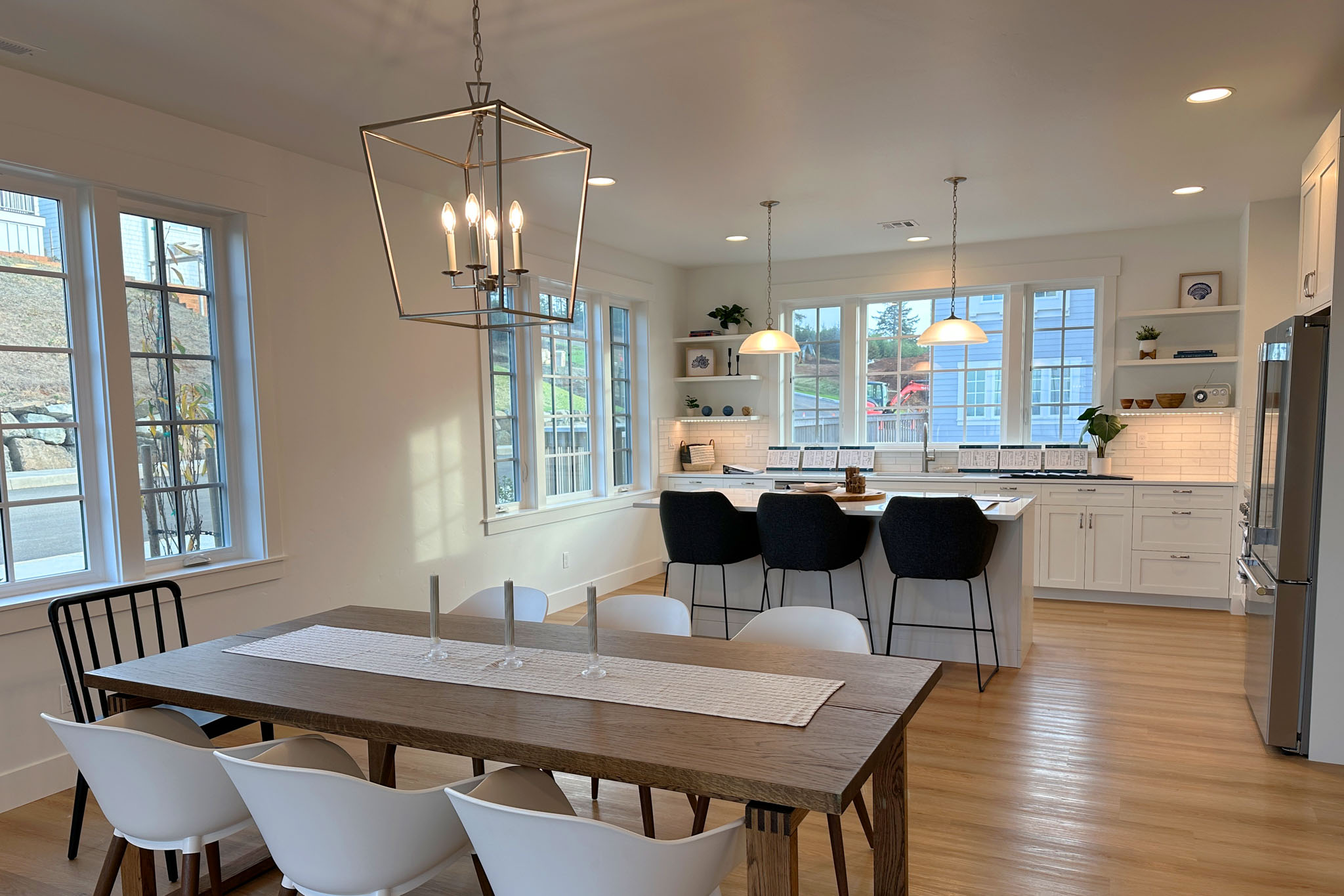 Modern open-concept kitchen and dining area with wooden floor, a large wooden dining table with white and black chairs, pendant lights over the kitchen island with black stools, white cabinetry, and large windows letting in natural light.