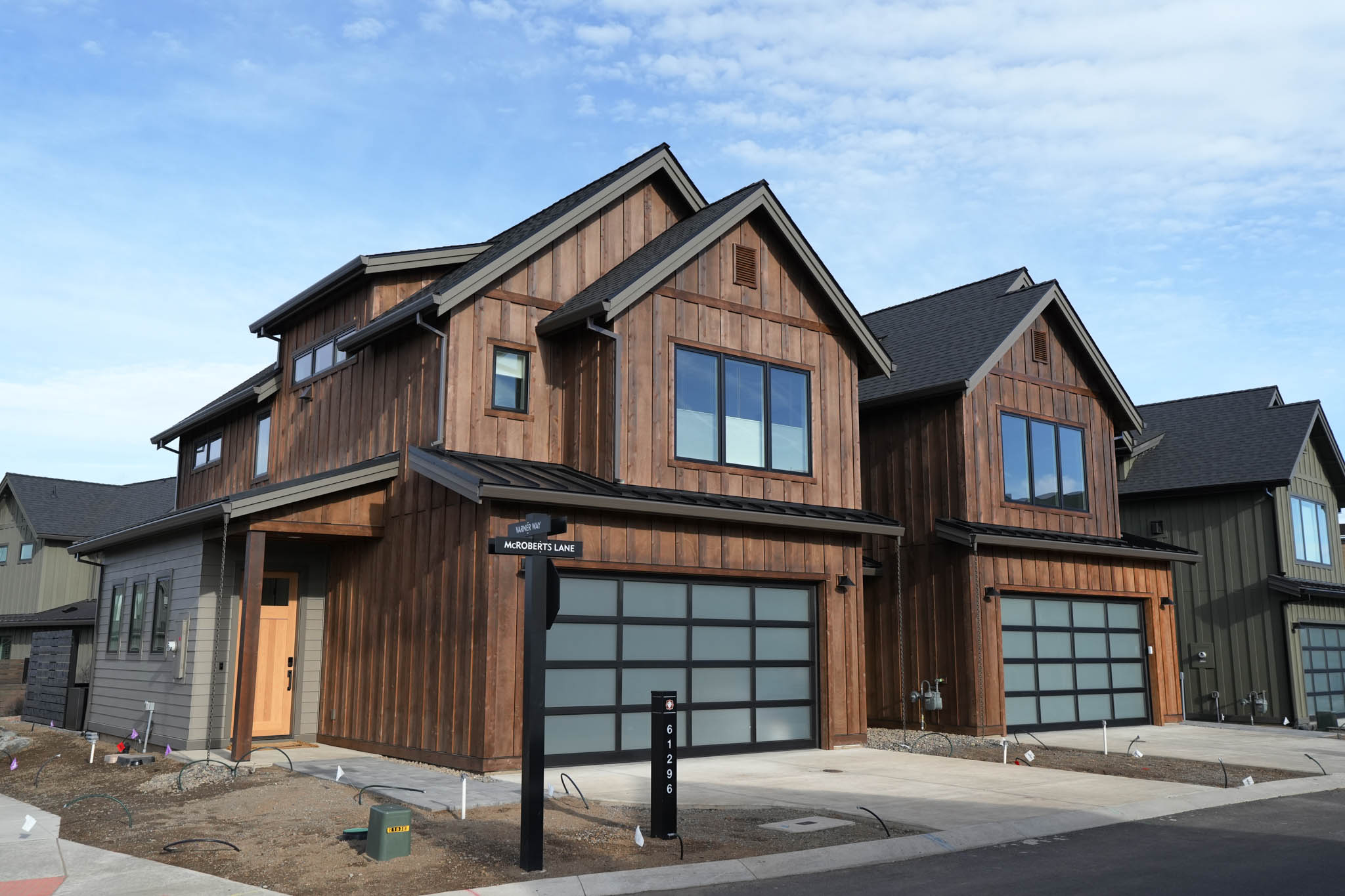 Street view of two modern two-story houses with wood-paneled exteriors, large frosted glass garage doors, and multiple windows under a blue sky.