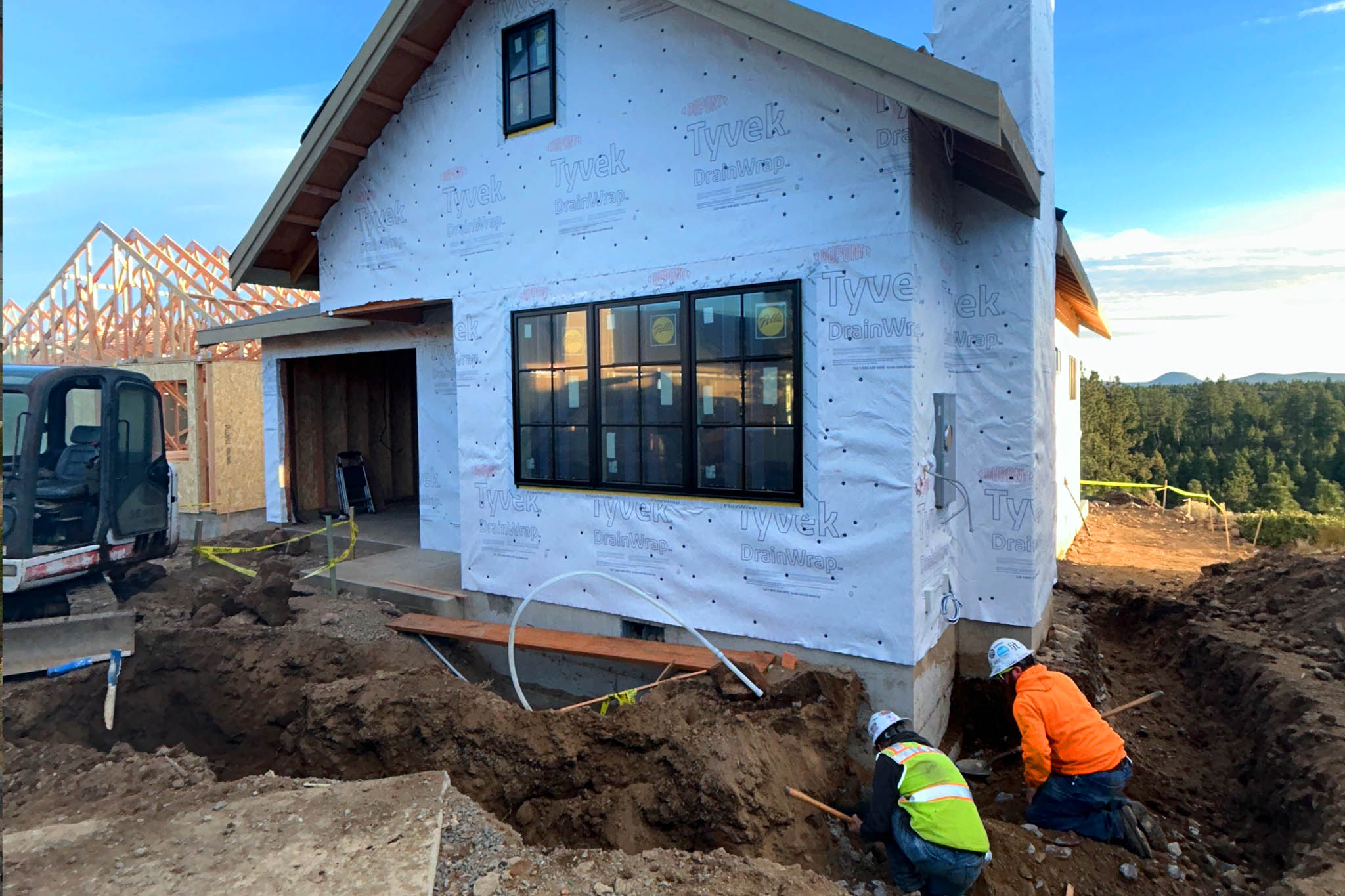 Two construction workers wearing hard hats and safety vests digging trenches near the foundation of a partially built house wrapped in white Tyvek DrainWrap, with visible window and garage framing, a small excavator, and forested hills in the background under a blue sky.