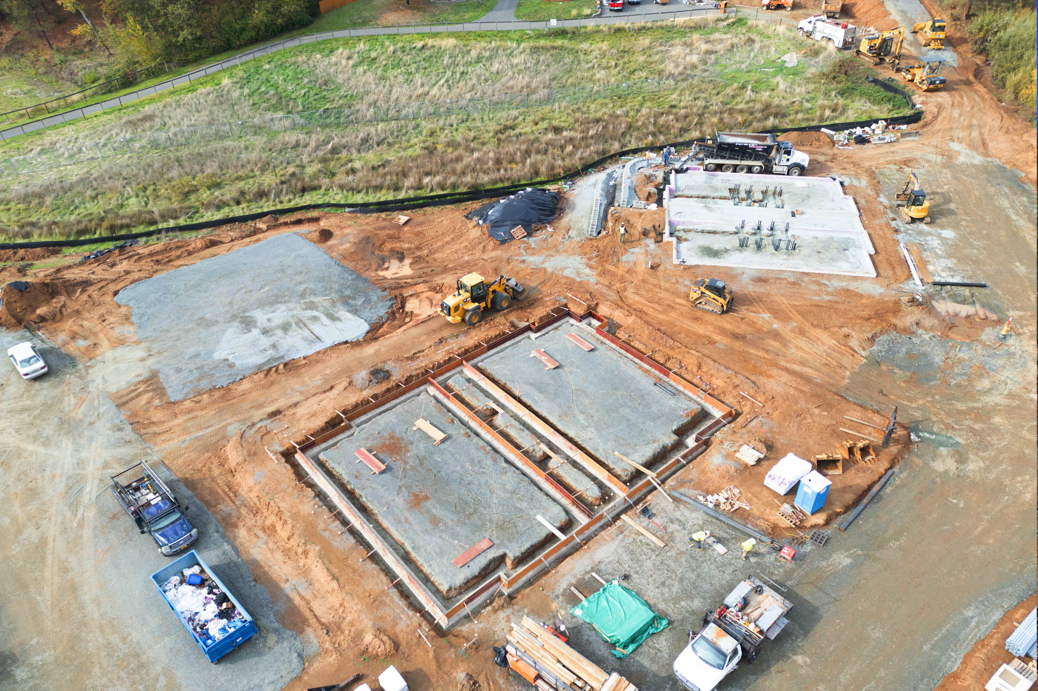 Aerial view of a construction site with building foundations in progress, construction vehicles, workers, and equipment on dirt terrain surrounded by grass.