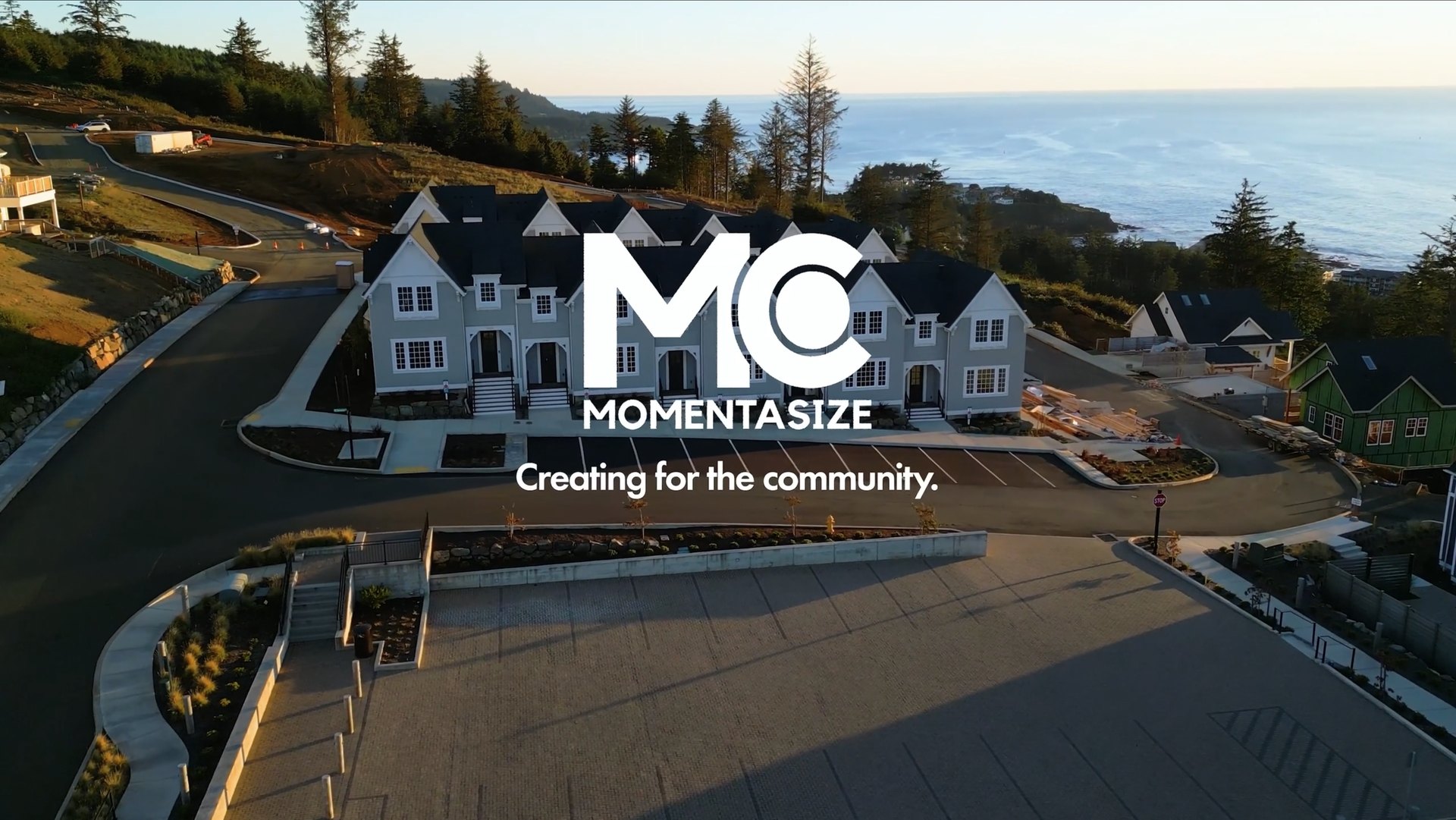 Aerial view of a residential neighborhood near a coastline at sunset with a row of gray and white houses, curved roads, trees, and ocean in the background, overlaid with white text: 'MC MOMENTASIZE Creating for the community.'