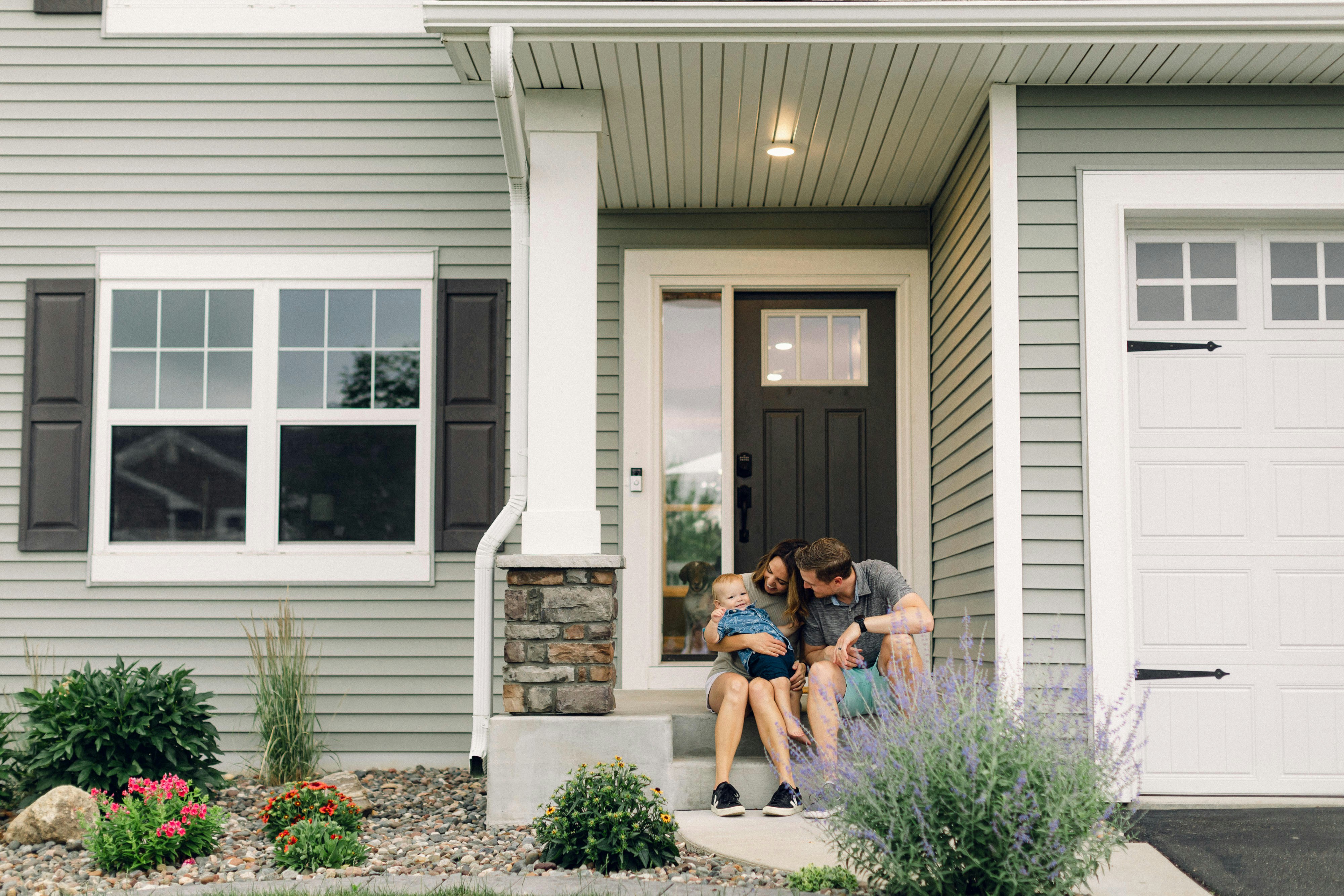 A family with a mother, father, and baby sitting on the front steps of a gray house with a black front door and white garage door, with a dog visible inside through the door window.