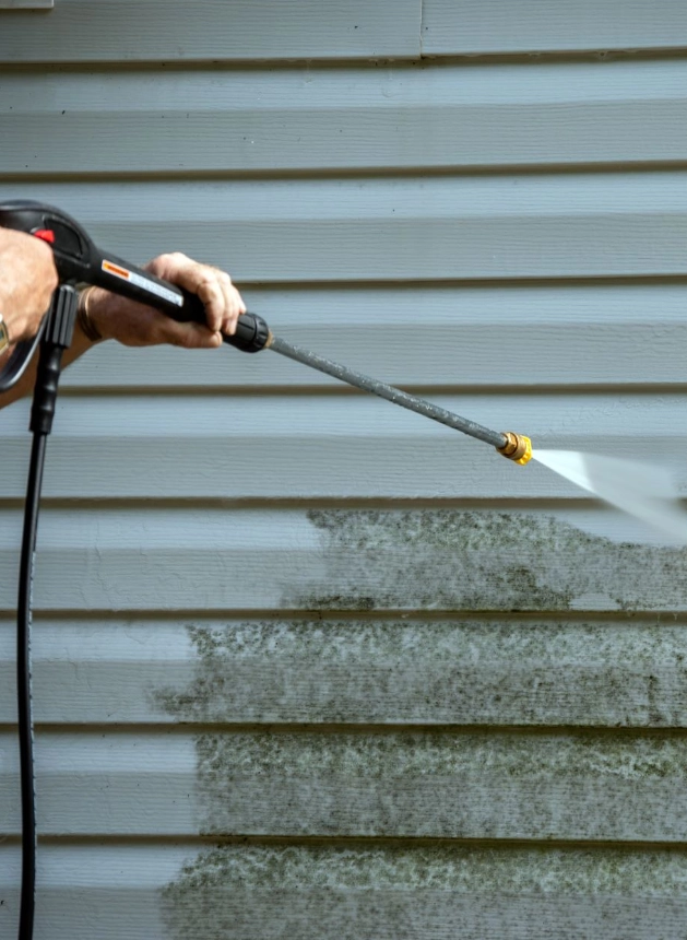 Person using a pressure washer to clean mold and dirt off the exterior siding of a house, showing a clear difference between cleaned and uncleaned areas.