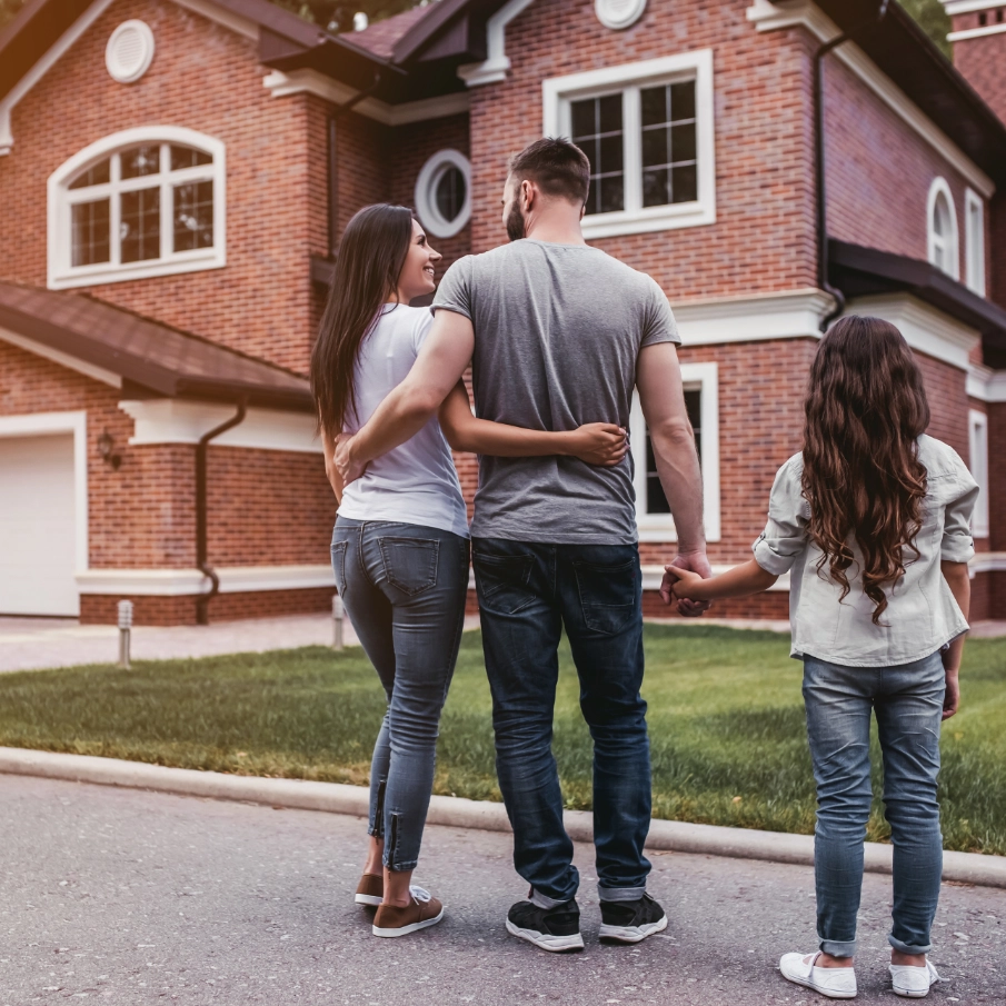 A family of three with a woman, man, and young girl holding hands and standing outside a large brick house with multiple windows.