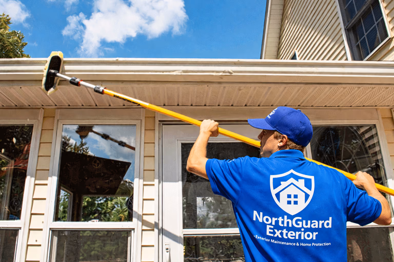 Man in a blue NorthGuard Exterior shirt and cap power washing the exterior siding and eaves of a beige house under a partly cloudy sky.