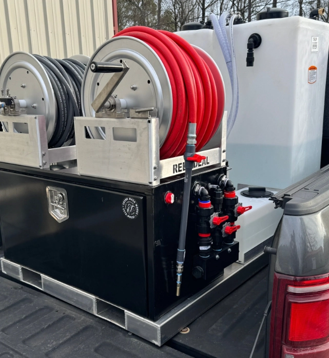 Truck bed equipped with a black skid unit featuring two hose reels, one with a red hose and one with a gray hose, attached to a large white liquid storage tank with plumbing valves and connectors.