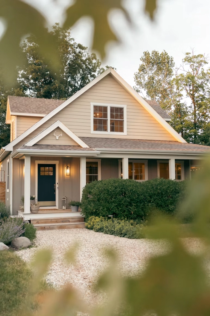 Two-story house with beige siding and brown roof, featuring a front porch with white columns and a blue front door, surrounded by green bushes and trees in a peaceful yard.