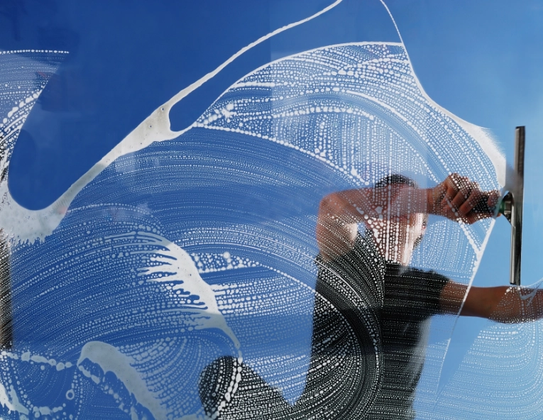 Person cleaning a large glass window with a squeegee, seen through the soapy suds on the glass against a clear blue sky.