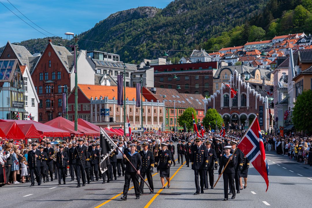 Et buekorps marsjerer gjennom Bergen sentrum, med publikum, byhus og fjellsiden i bakgrunnen.