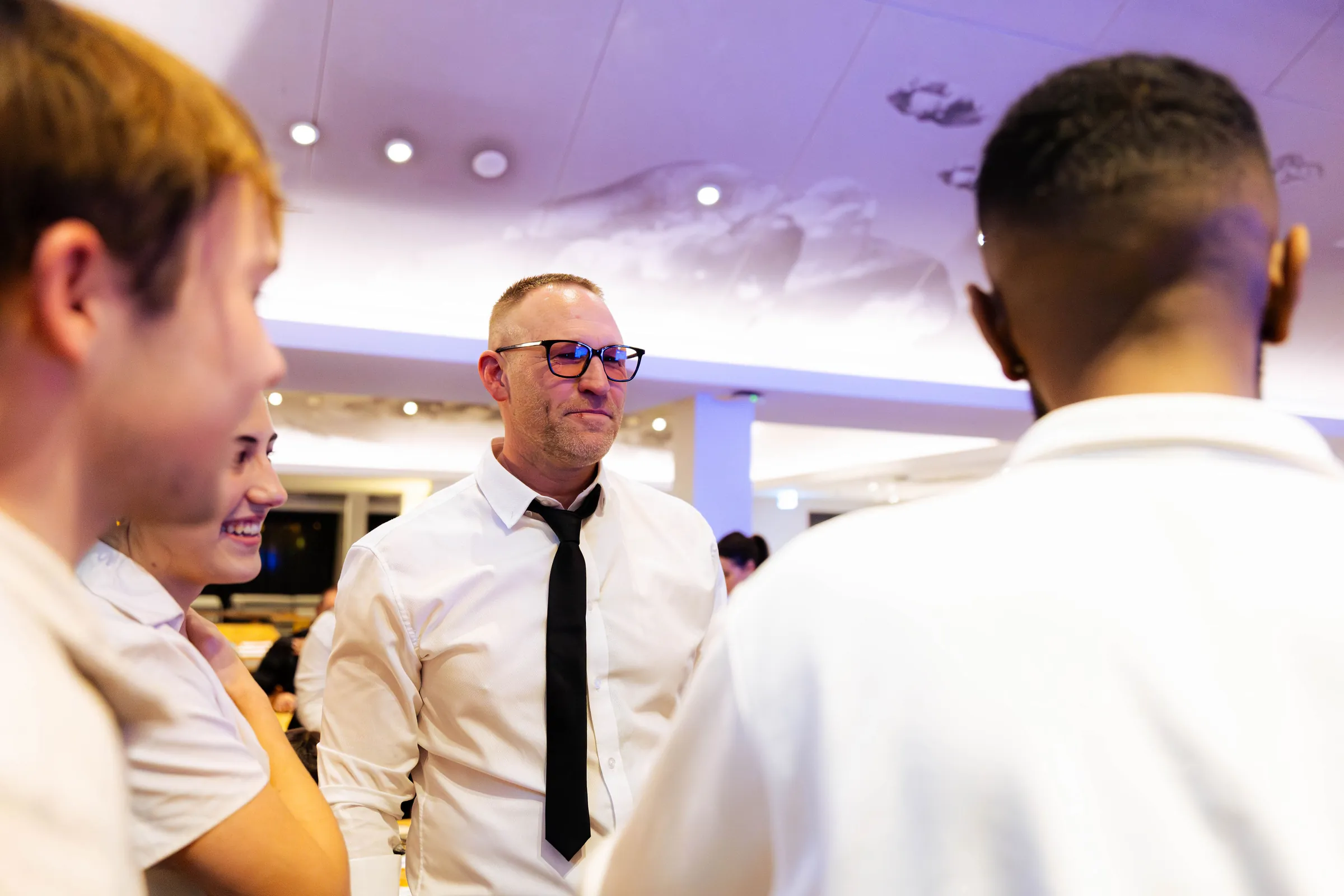 A guest in a white shirt and black tie talking with others at the Armed For Life quiz night at The Hall, The Camfield, Burswood.
