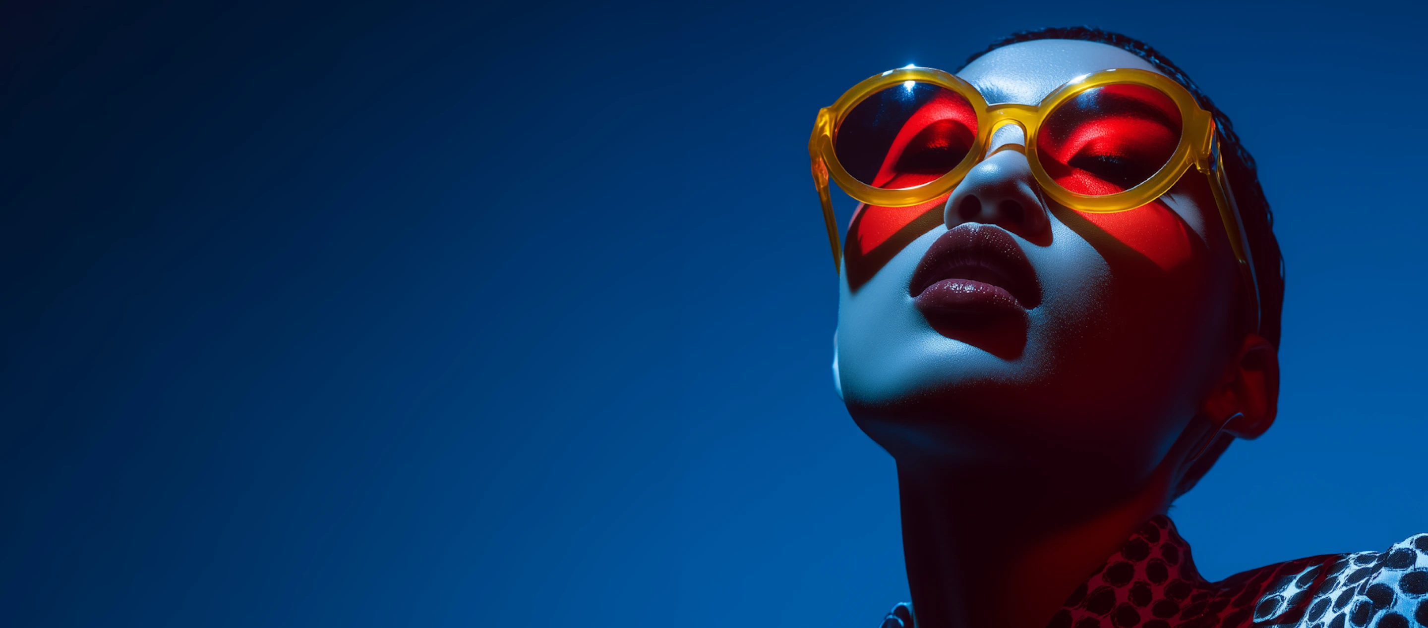 Close-up of a person wearing yellow-framed red sunglasses with dramatic blue and red lighting on their face against a dark blue background.